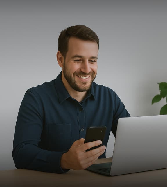 Man smiling while using his phone and laptop.